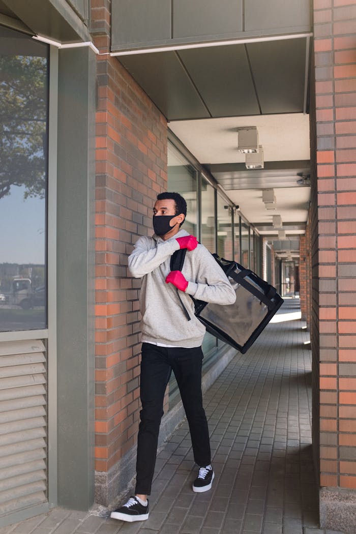 Courier in a face mask and gloves delivers food outdoors, walking by brick buildings.